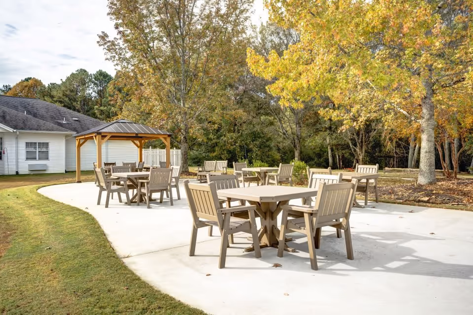 Outdoor patio area with multiple beige tables and chairs on a concrete surface, surrounded by green grass and trees with autumn foliage. A small wooden gazebo is visible near a white building in the background.