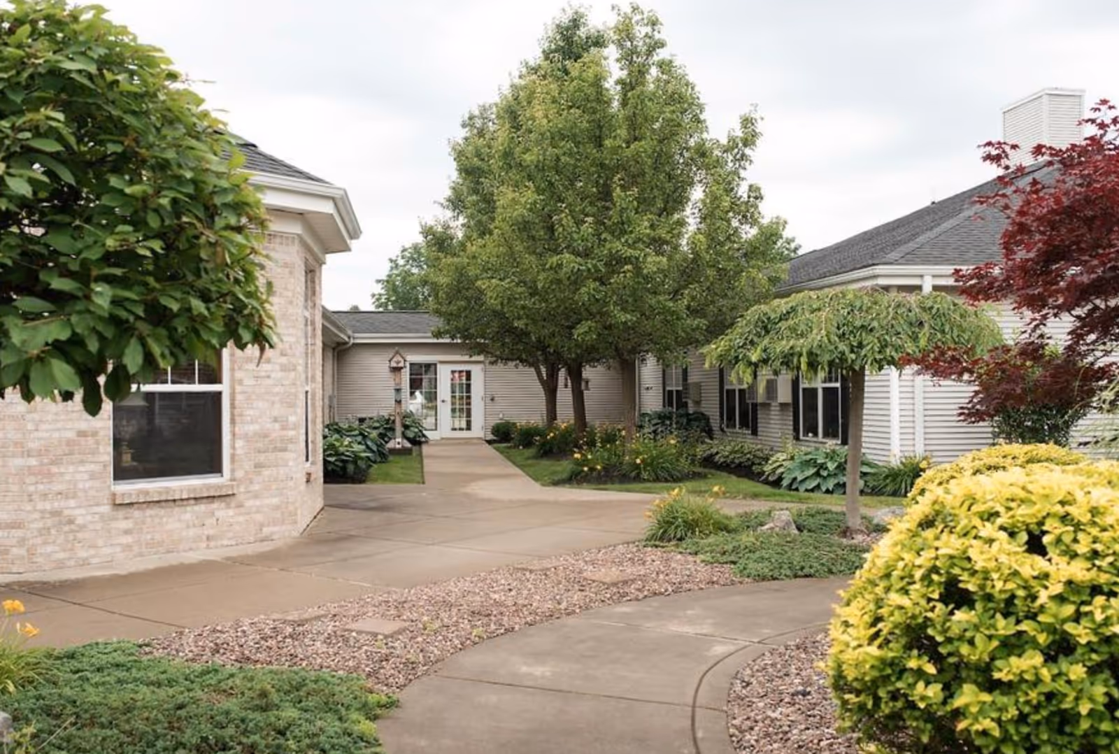 Landscaped courtyard and curved walkway leading to the entrance of a single-story senior living building with trees and shrubs.