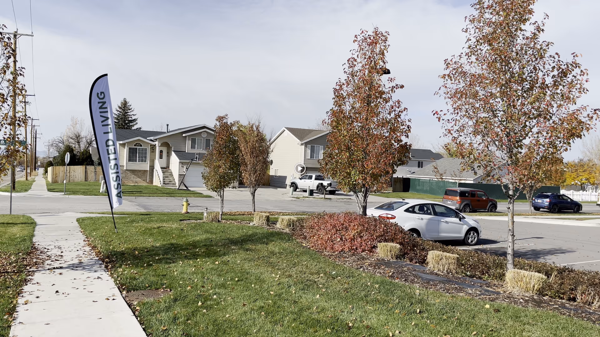 Outdoor view of a residential area with a sidewalk, green grass, small trees with autumn leaves, and a parking lot with several parked cars. A tall vertical flag with the text 'ASSISTED LIVING' is placed near the sidewalk.