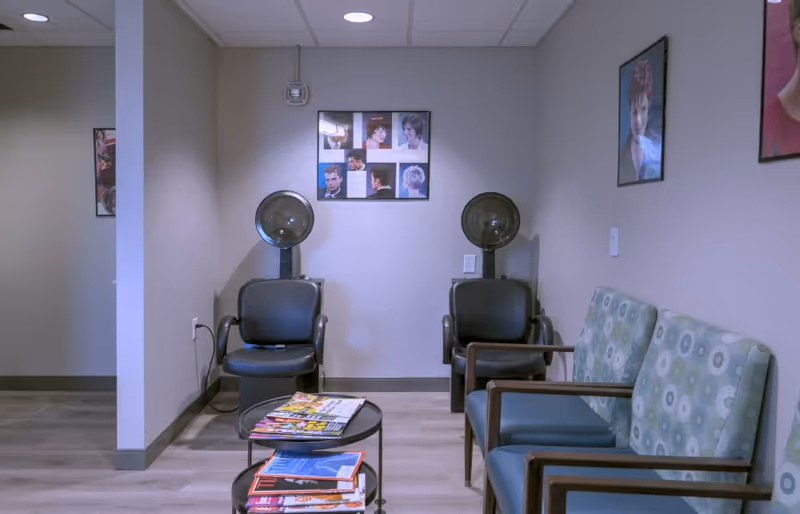 A small salon area with two black salon chairs equipped with hair dryers against a light gray wall. There are framed pictures of various hairstyles on the walls. To the right, there are three cushioned chairs with patterned upholstery. In front of the chairs is a small round table with magazines on it. The floor is light wood, and the ceiling has recessed lighting.