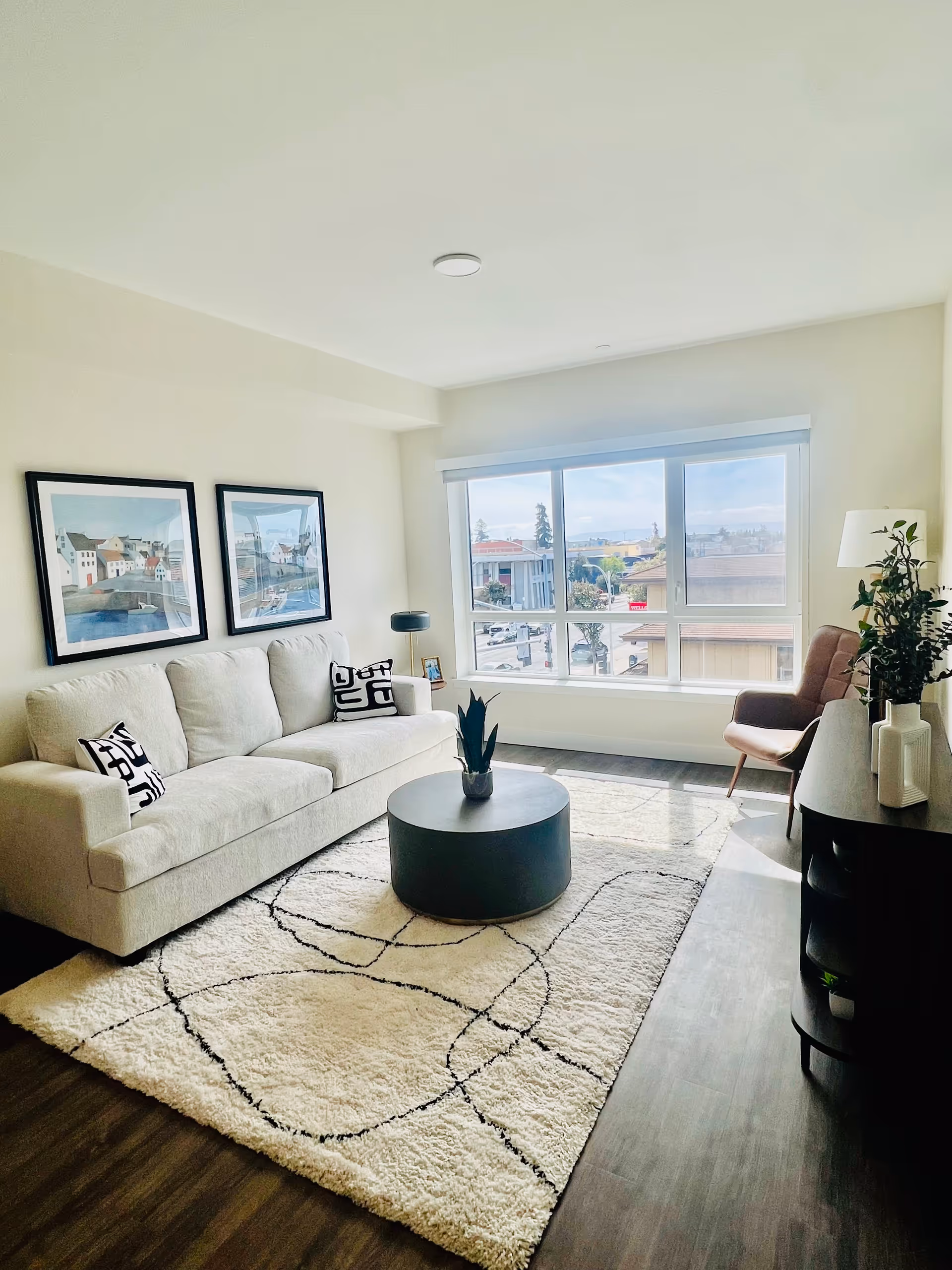 A bright living room with a large window letting in natural light. The room features a beige sofa with patterned cushions, a round black coffee table with a small plant on it, a patterned white rug, a brown armchair, a floor lamp, and a dark wooden side table with decorative vases and a plant. Two framed pictures hang on the wall above the sofa.