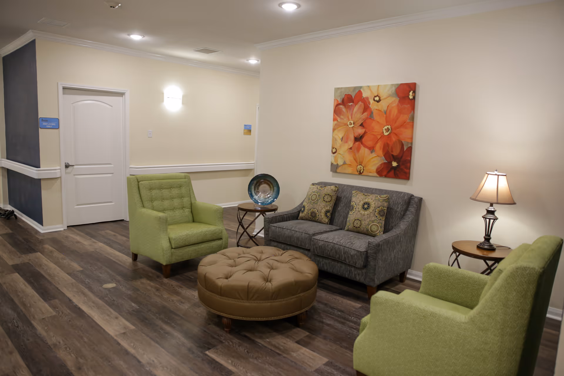 Small lounge with green armchairs, a gray sofa, a round tufted ottoman, side tables, a lamp and floral wall art.