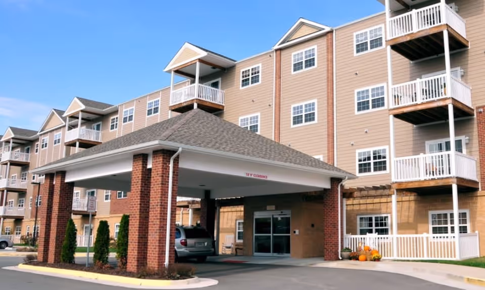 Front entrance of a multi-story senior living building with a covered porte-cochere, balconies, and parked car.