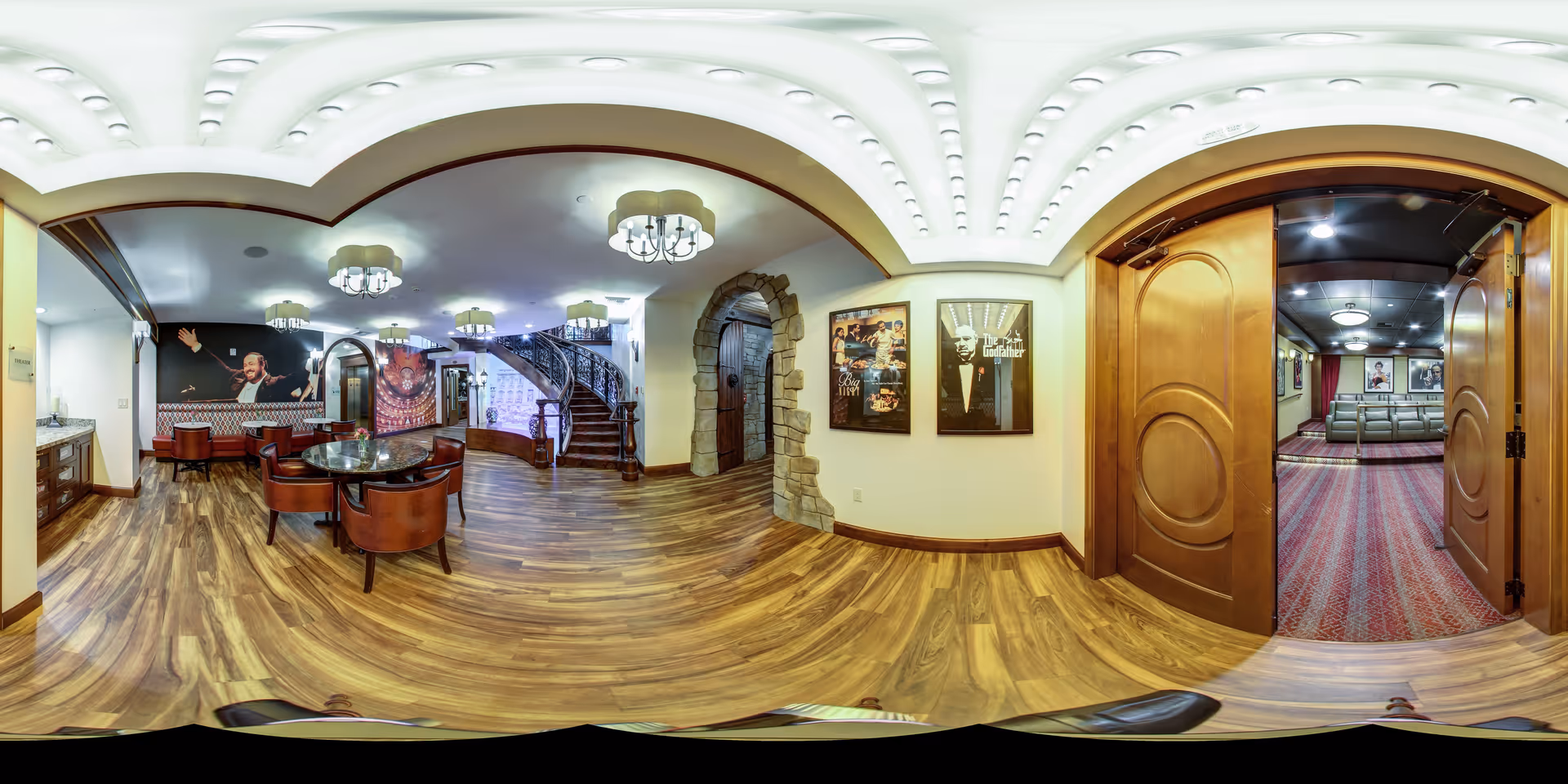 Panoramic interior of a senior living common area with seating around a round table, decorative posters, a staircase, and open double doors leading to a theater room.