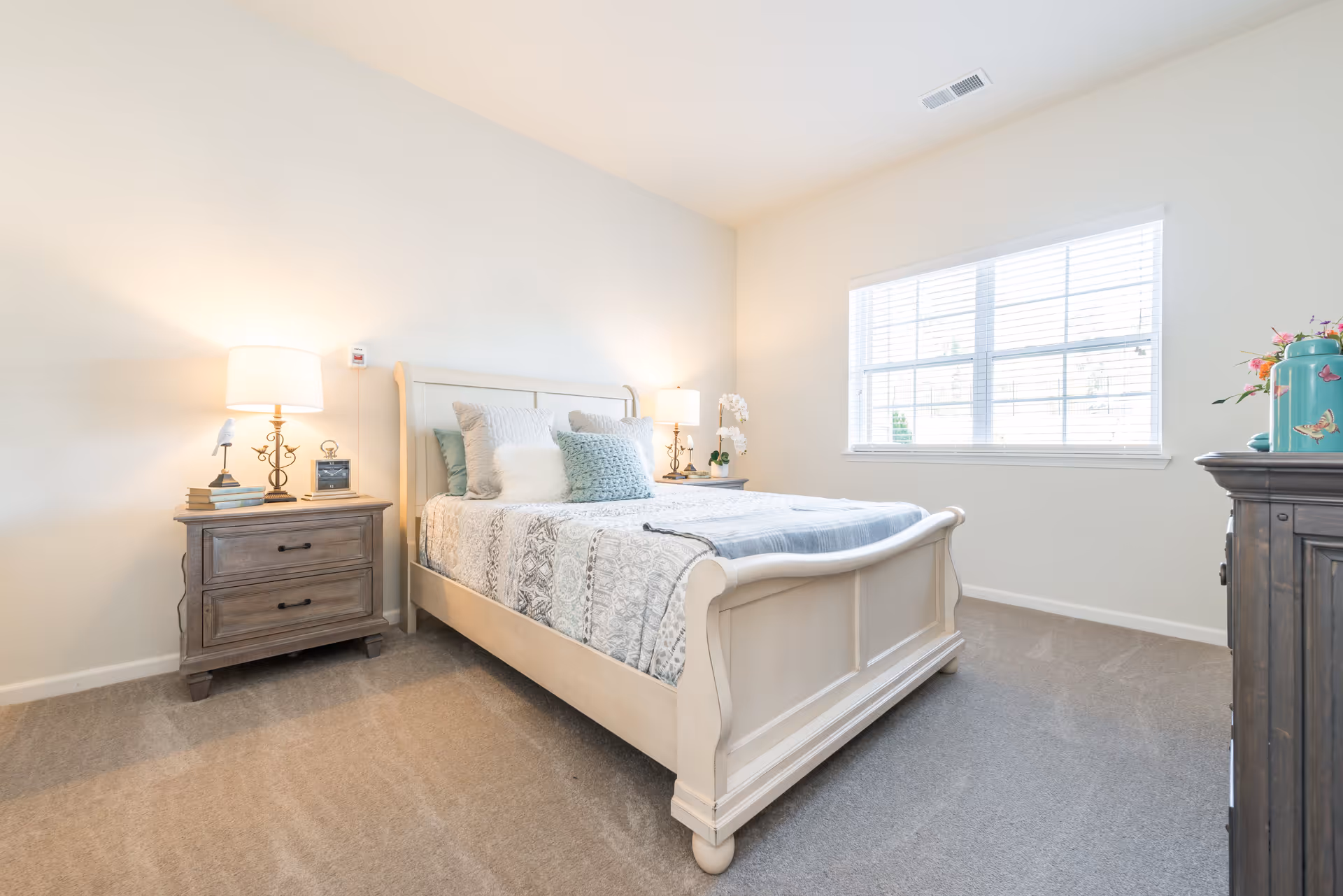 A bright and cozy bedroom with a white wooden bed frame, a neatly made bed with patterned bedding and multiple pillows. There are two wooden nightstands on either side of the bed, each with a lamp and decorative items. A large window with white blinds allows natural light to fill the room. The carpeted floor and light-colored walls create a warm and inviting atmosphere.