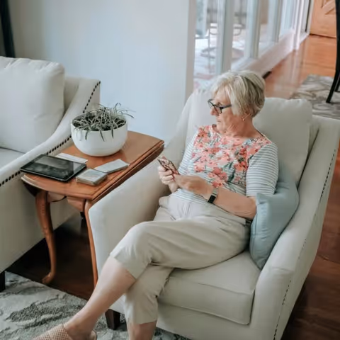 An elderly woman with short gray hair and glasses sits comfortably in a beige armchair in a well-lit living room. She is wearing a floral patterned top with striped sleeves and light-colored pants, looking at her smartphone. Next to her is a wooden side table with a white potted plant, a tablet, and some cards. The room has wooden flooring and a patterned rug, with another beige armchair partially visible.