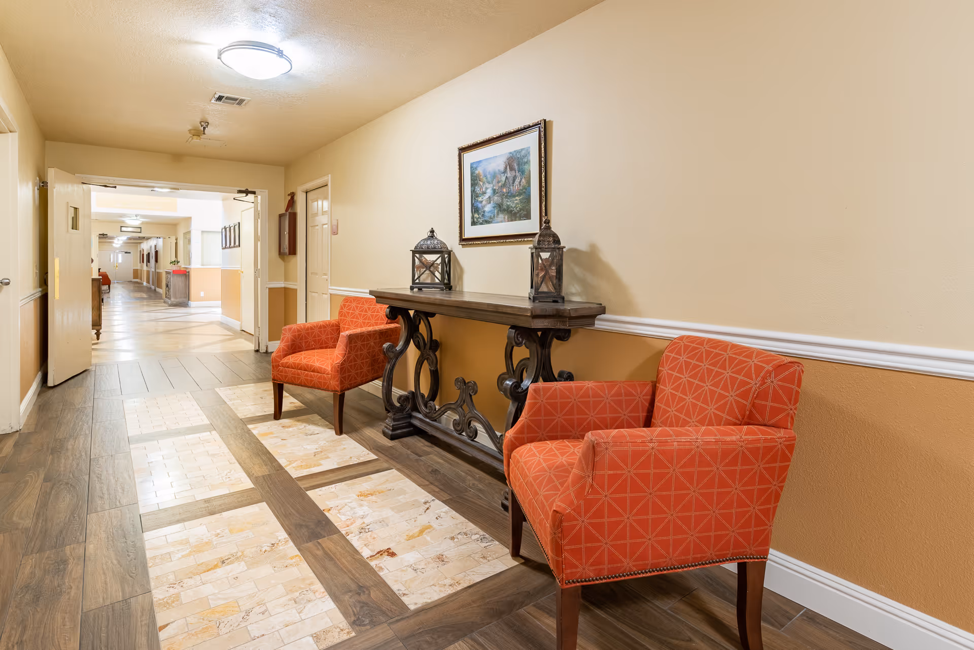 Well-lit hallway seating area with two orange patterned chairs, a decorative console table with lanterns, and framed art along a corridor.