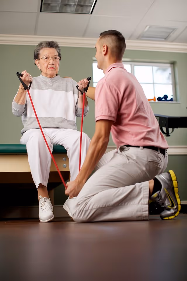 An elderly woman sitting on a bench exercising with red resistance bands while a young male therapist kneels in front of her, assisting with the exercise in a well-lit room with green walls and a window in the background.