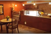 Interior view of a dining area with a round table and chairs in the foreground, and a food service counter with a glass sneeze guard in the background. The room has warm lighting, decorative flowers on the table and counter, and a tiled floor.