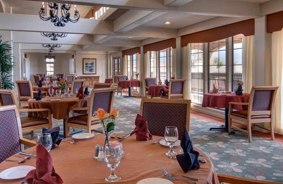Dining room with multiple round tables set with glassware and folded napkins, chairs, chandeliers, and large windows letting in natural light.