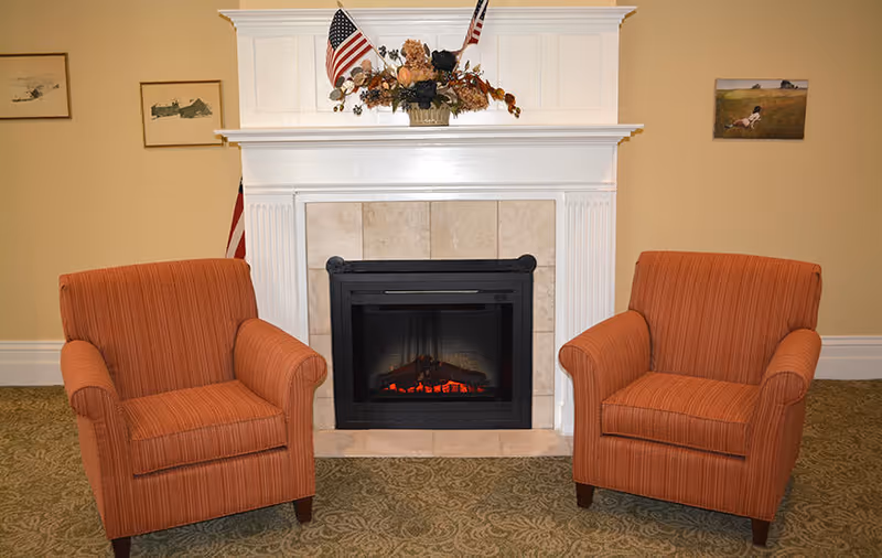 Two orange upholstered armchairs flanking a tiled fireplace with a white mantel decorated with flags and a floral arrangement.