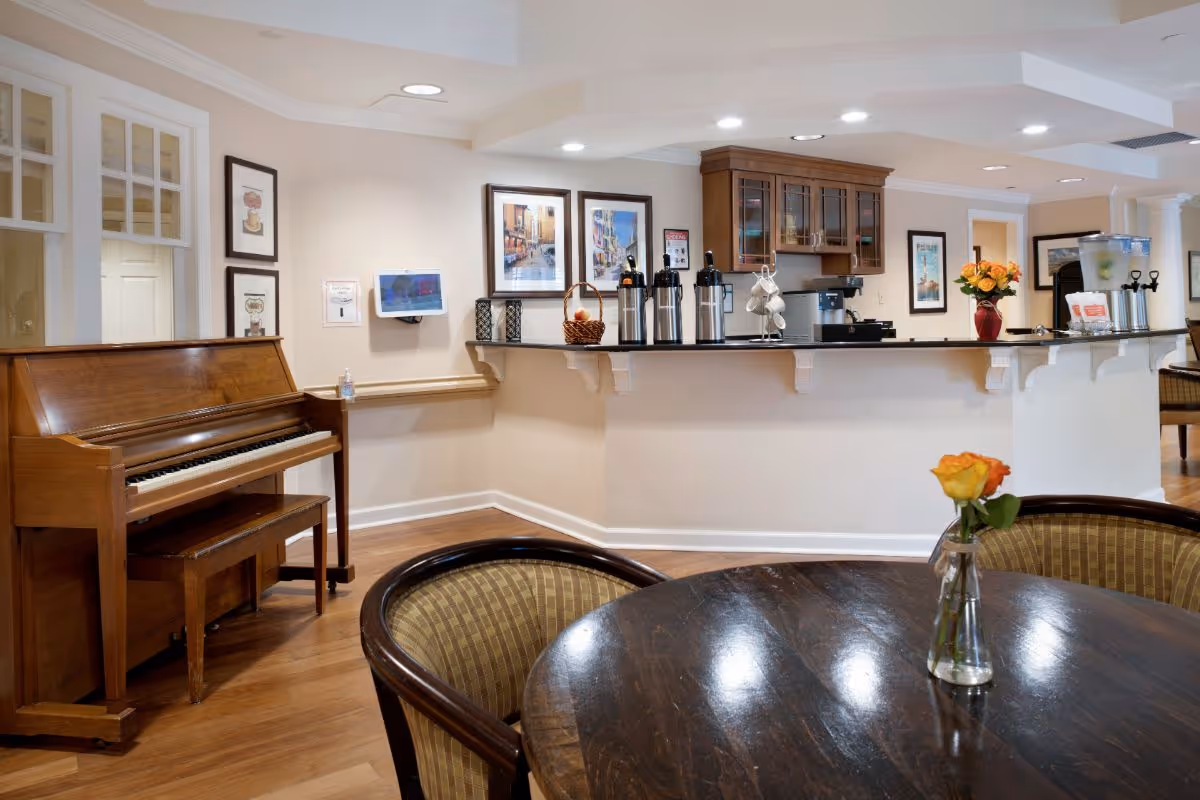 Bright communal dining/lounge area with a piano, a serving counter with beverage dispensers, and a round table holding a single rose in a vase.