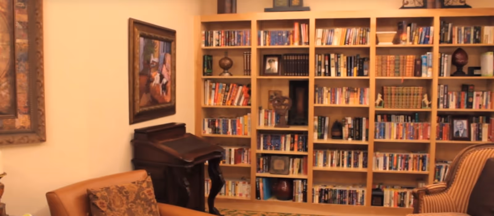 A cozy interior room featuring a large wooden bookshelf filled with books and decorative items. There is a wooden antique desk with curved legs against the wall, a framed painting above it, and two upholstered chairs with patterned cushions in the foreground.