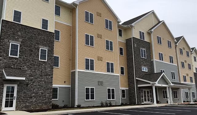 Exterior view of a multi-story senior living facility building with beige, gray, and stone facade, multiple windows, and a covered entrance. The parking lot is visible in the foreground.