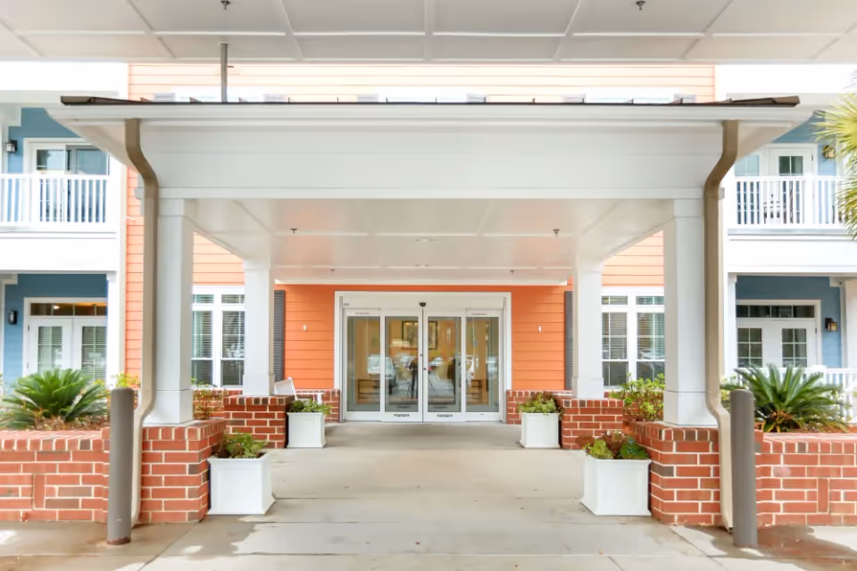 Entrance to a senior living facility with a covered driveway supported by white columns, brick planters with green plants, and double glass doors leading inside. The building exterior features orange and blue siding with white trim and balconies.