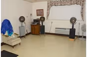 A room with three vintage hair dryers on stands, a beige armchair with a blue cloth draped over it, a wooden cabinet with a stereo system, a framed picture on the wall, a window with a valance, and a beige tiled floor.
