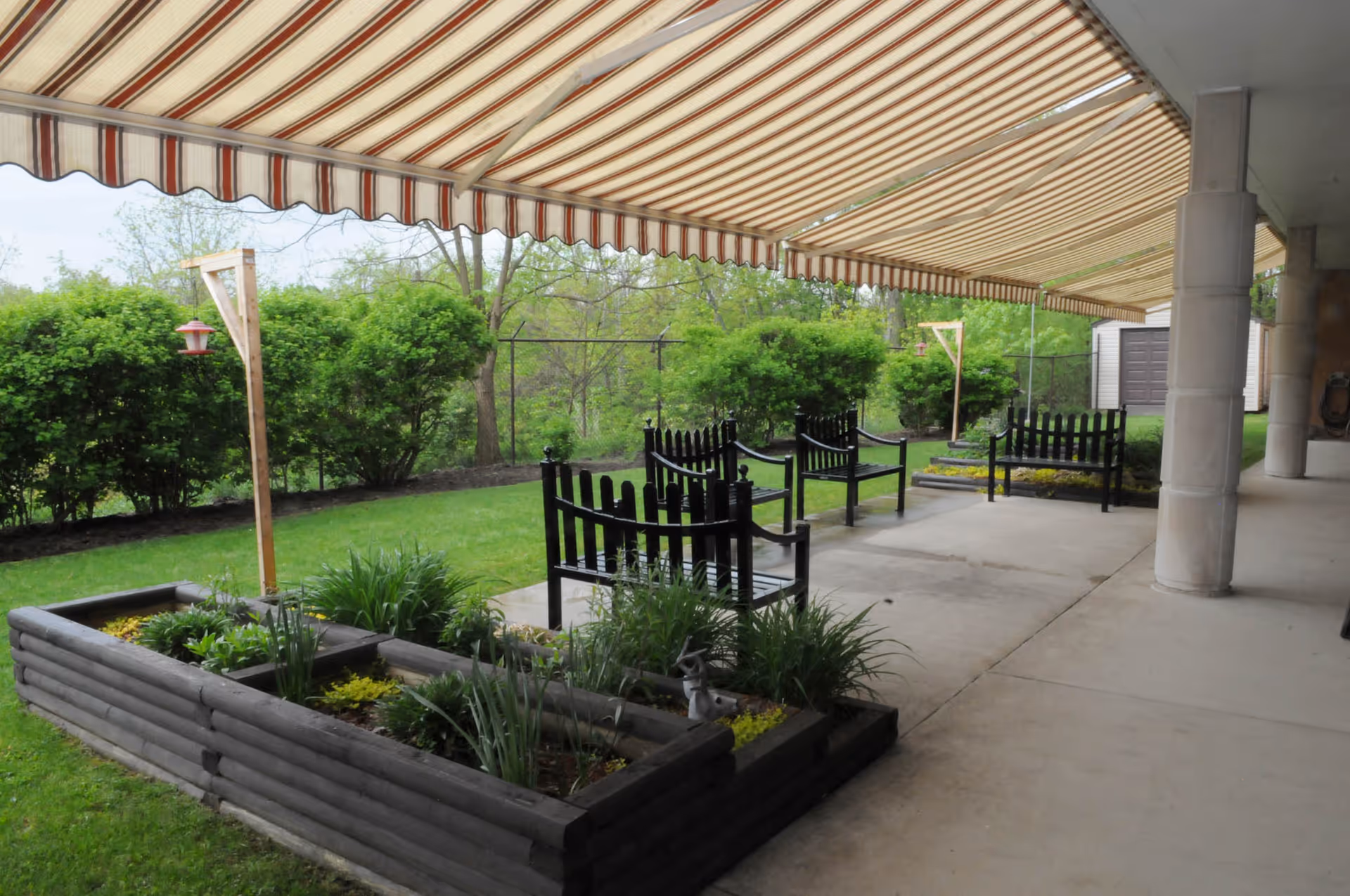 Covered outdoor patio area with a striped awning, black wooden benches, raised garden beds with plants, and green bushes in the background.