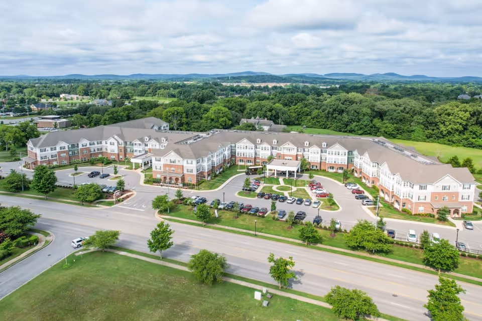 Aerial view of a large senior living facility named Harmony at Victory Station, surrounded by greenery and parking lots, with a road in the foreground and hills in the distance under a cloudy sky.