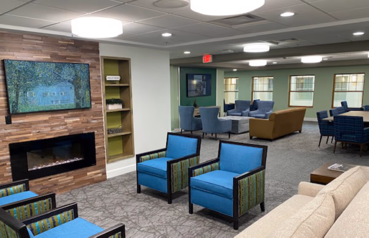 A spacious senior living common area with multiple seating arrangements including blue armchairs, a beige sofa, and a brown couch. The room features a modern electric fireplace set in a wooden accent wall with a large flat-screen TV mounted above it. There are several windows letting in natural light, and the ceiling has recessed lighting and round light fixtures.