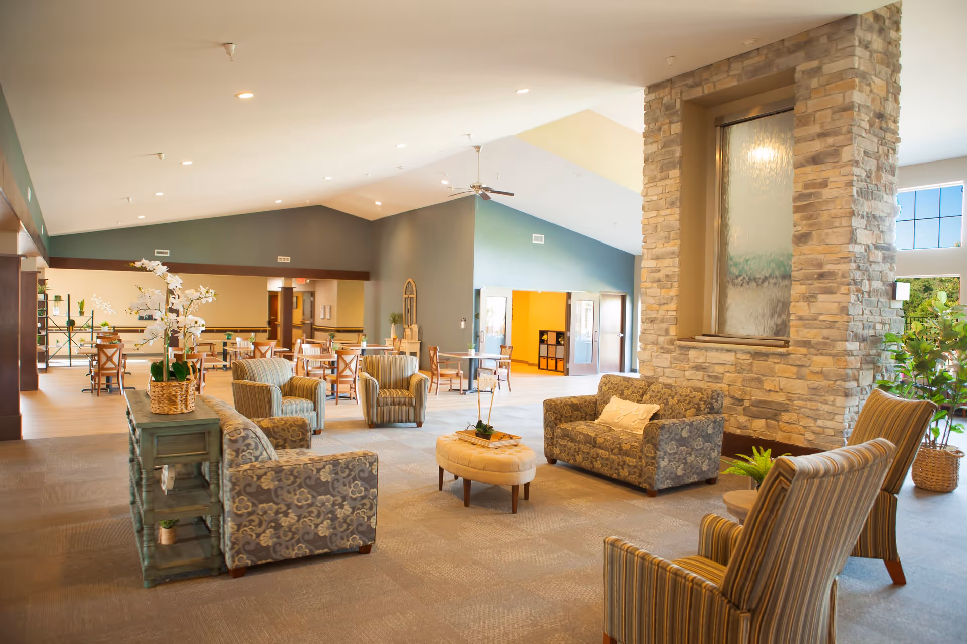 A spacious and well-lit common area in a senior living facility featuring comfortable patterned armchairs and sofas arranged around a central ottoman. The room has a high ceiling with recessed lighting and a ceiling fan. There is a large stone fireplace with a water feature, and several tables and chairs are visible in the background near a green accent wall. Potted plants and decorative items add a welcoming touch.