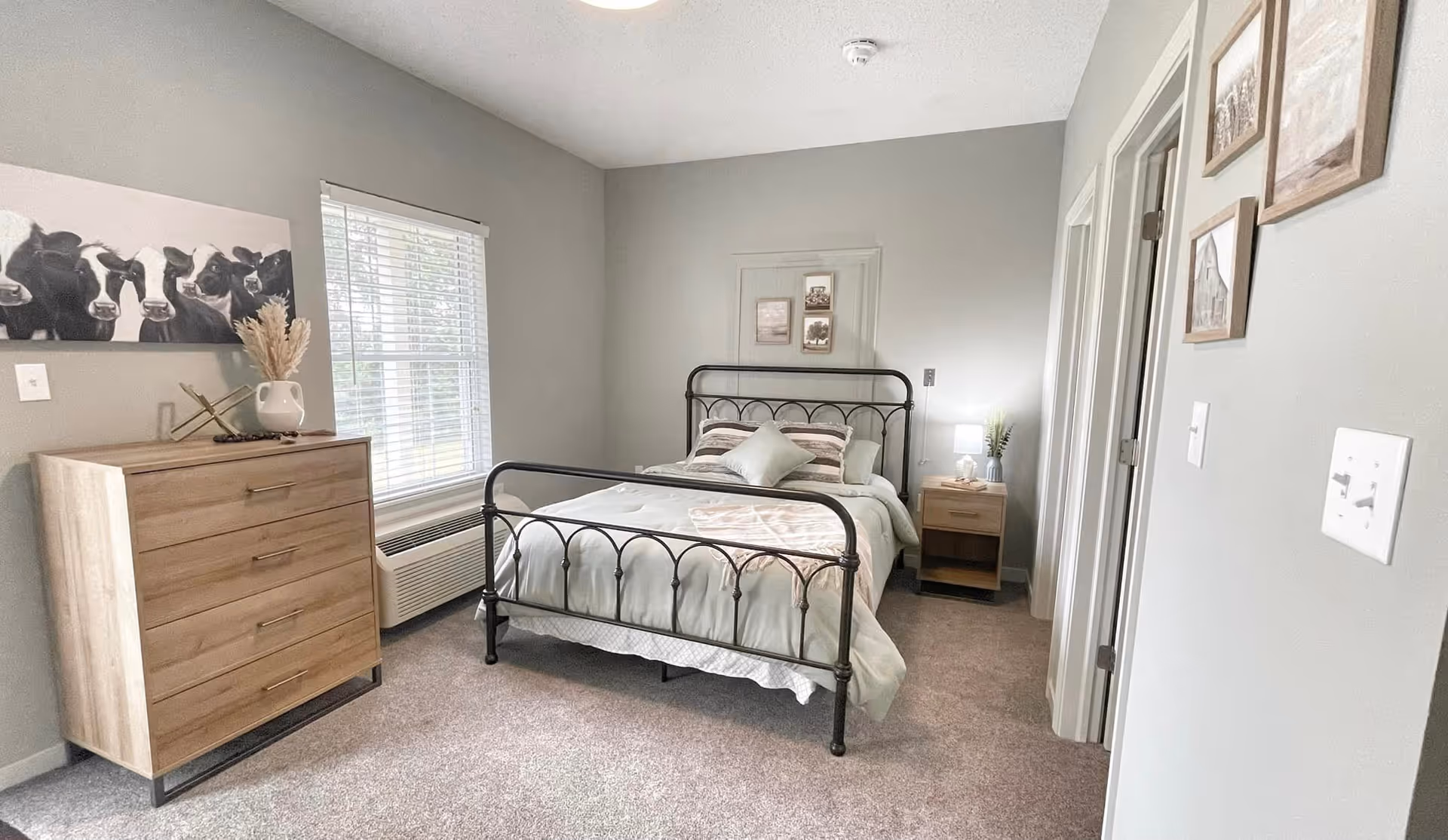 A bedroom with a metal frame bed made with light-colored bedding and pillows. To the left of the bed is a wooden dresser with a decorative vase and artwork of cows above it. A window with blinds is next to the dresser. To the right of the bed is a wooden nightstand with a lamp and a small plant. The walls are painted light gray and there are framed pictures hanging on the wall near the door.