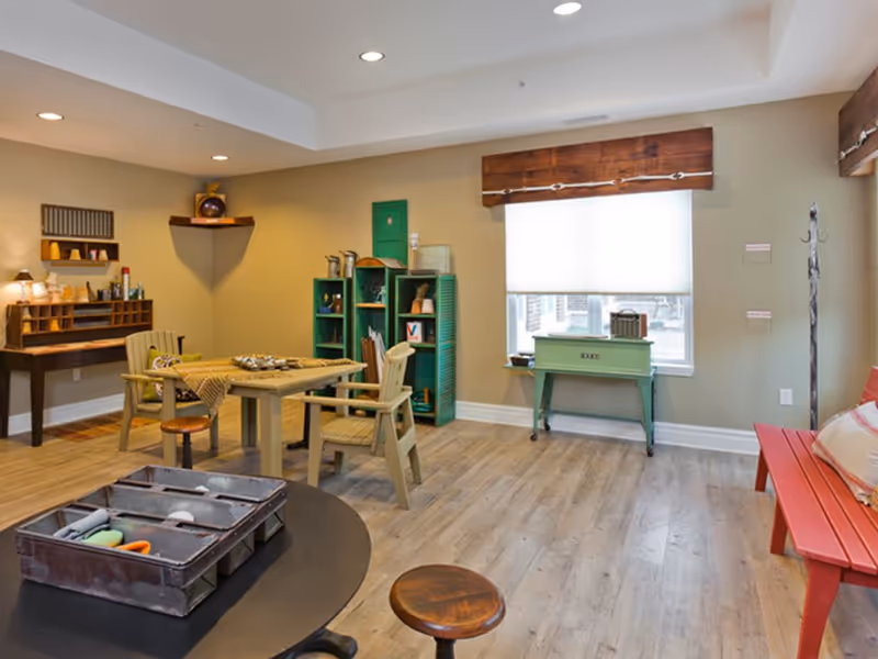 Well-lit communal activity room with tables, chairs, shelving and a window with a wooden valance.