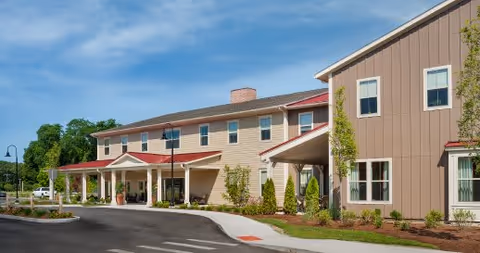 Exterior view of Carriage House at Lee's Farm showing a two-story building with beige and brown siding, multiple windows, a covered entrance with columns, landscaped greenery, and a paved driveway under a clear blue sky.
