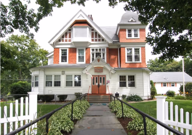 Front view of a large Victorian-style house with red shingled upper siding and white lower siding, a central entrance reached by steps and a walkway flanked by plants and white picket fencing.
