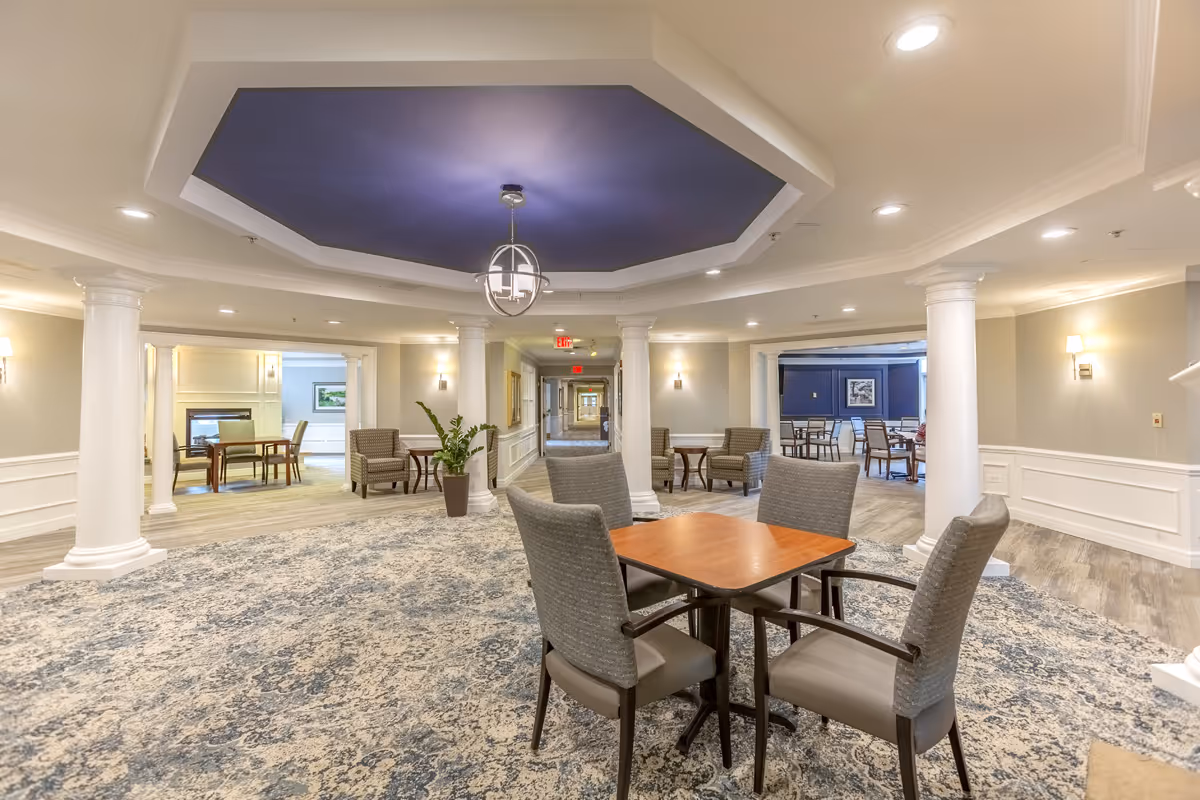 Spacious, well-lit common seating area with tables and chairs, columns, patterned carpet, and a blue recessed ceiling feature.