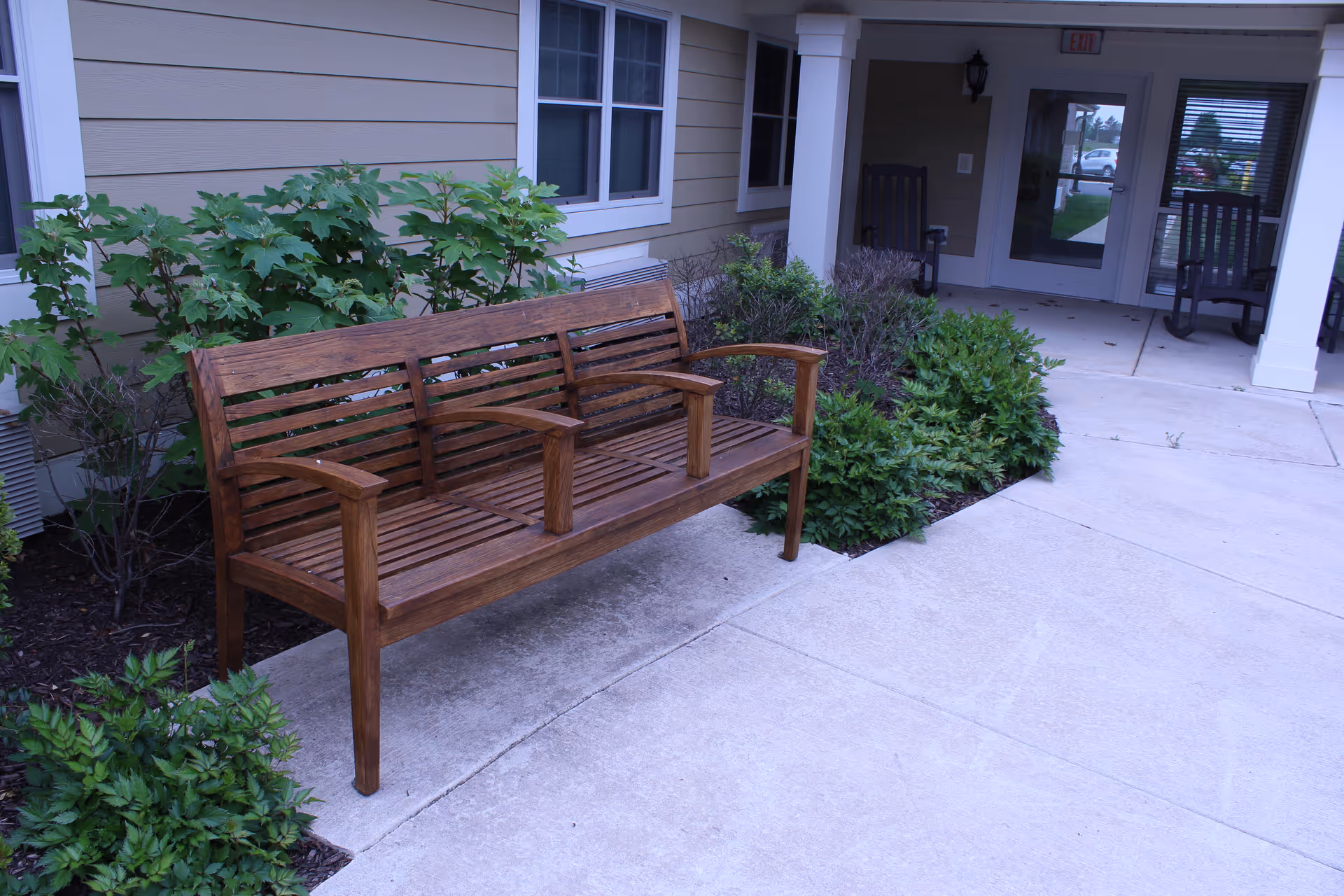 Wooden bench with armrests placed on a concrete walkway outside a building entrance, surrounded by green shrubs and plants, with two rocking chairs visible on the covered porch area.