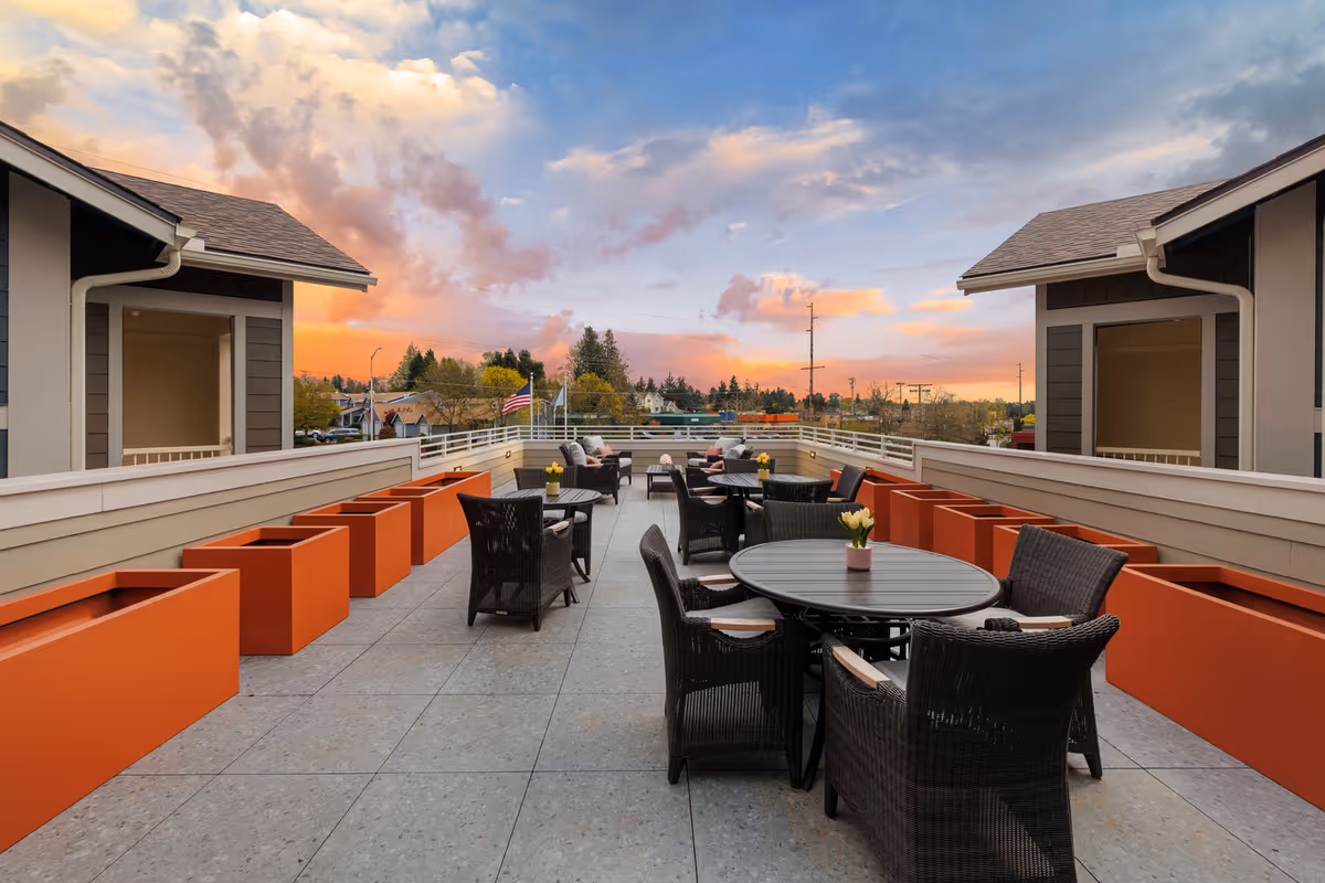 Outdoor patio area at sunset with round tables and wicker chairs arranged for seating. Orange planter boxes line the sides, and the sky is partly cloudy with warm colors from the setting sun. Buildings and trees are visible in the background.