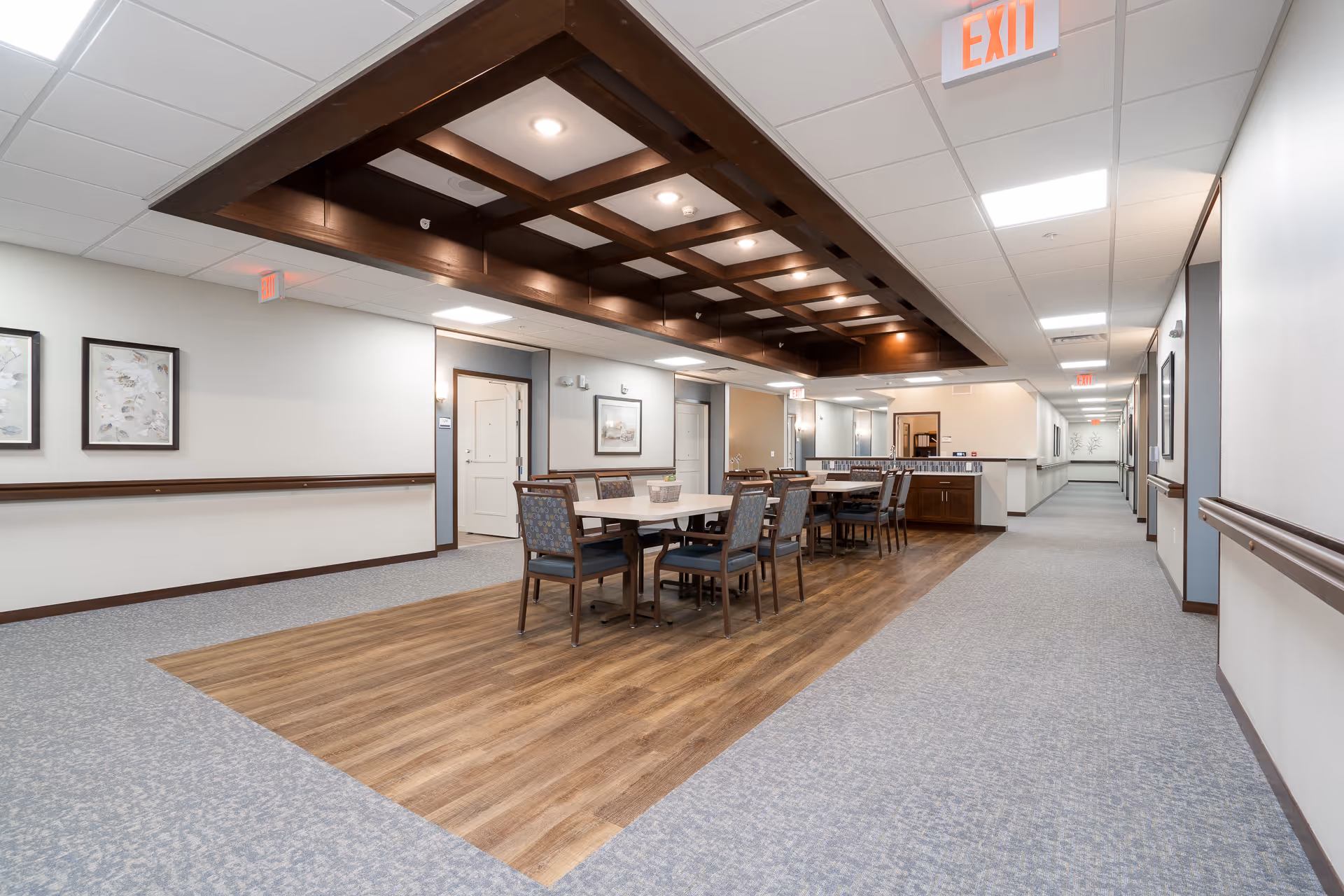 Interior view of a senior living facility hallway with a seating area in the center featuring two tables and multiple chairs. The floor has a wood section under the seating area and carpeted sections along the hallway. The ceiling has recessed lighting with a wooden grid design above the seating area. Walls are light-colored with framed artwork and handrails along the hallway. Exit signs are visible on the ceiling.