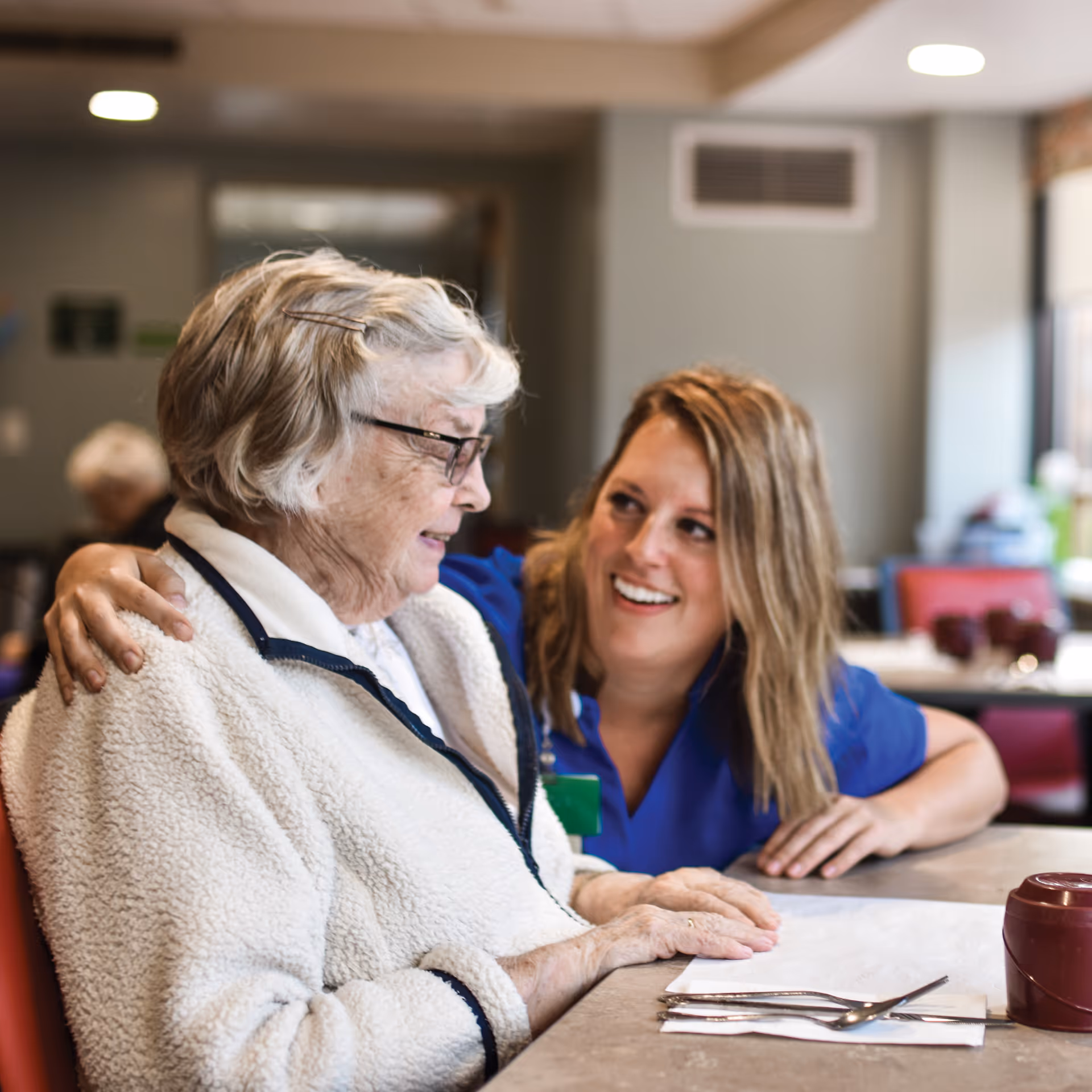 A smiling caregiver leans toward and embraces an elderly woman as they sit together at a dining table in a senior living facility.