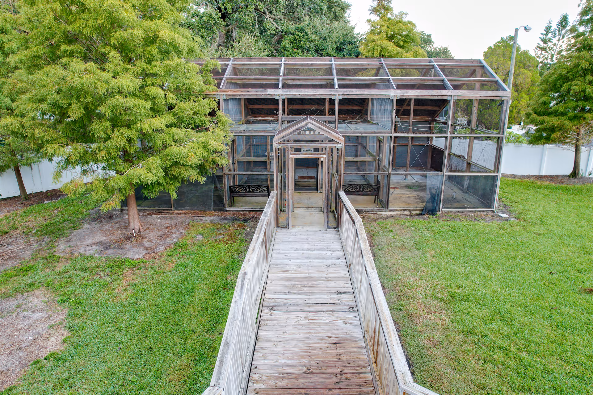 Wooden ramp leading to a large screened outdoor enclosure in a grassy yard with trees.