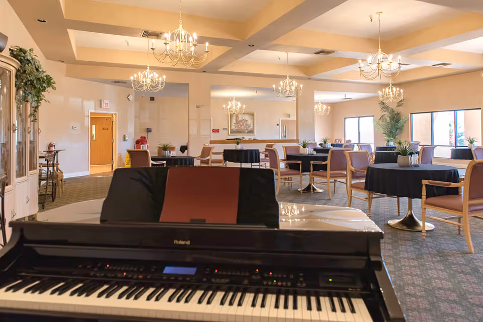 A communal dining/activity room with a piano in the foreground, round tables with black tablecloths, chairs, and chandeliers.