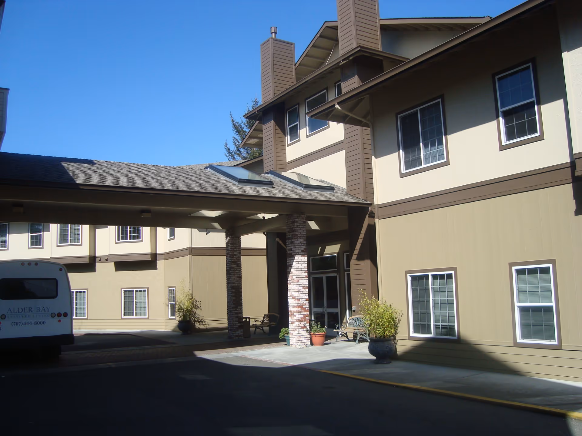 Exterior front entrance of the Alder Bay assisted living building with a covered portico, potted plants, benches, and a parked shuttle van.