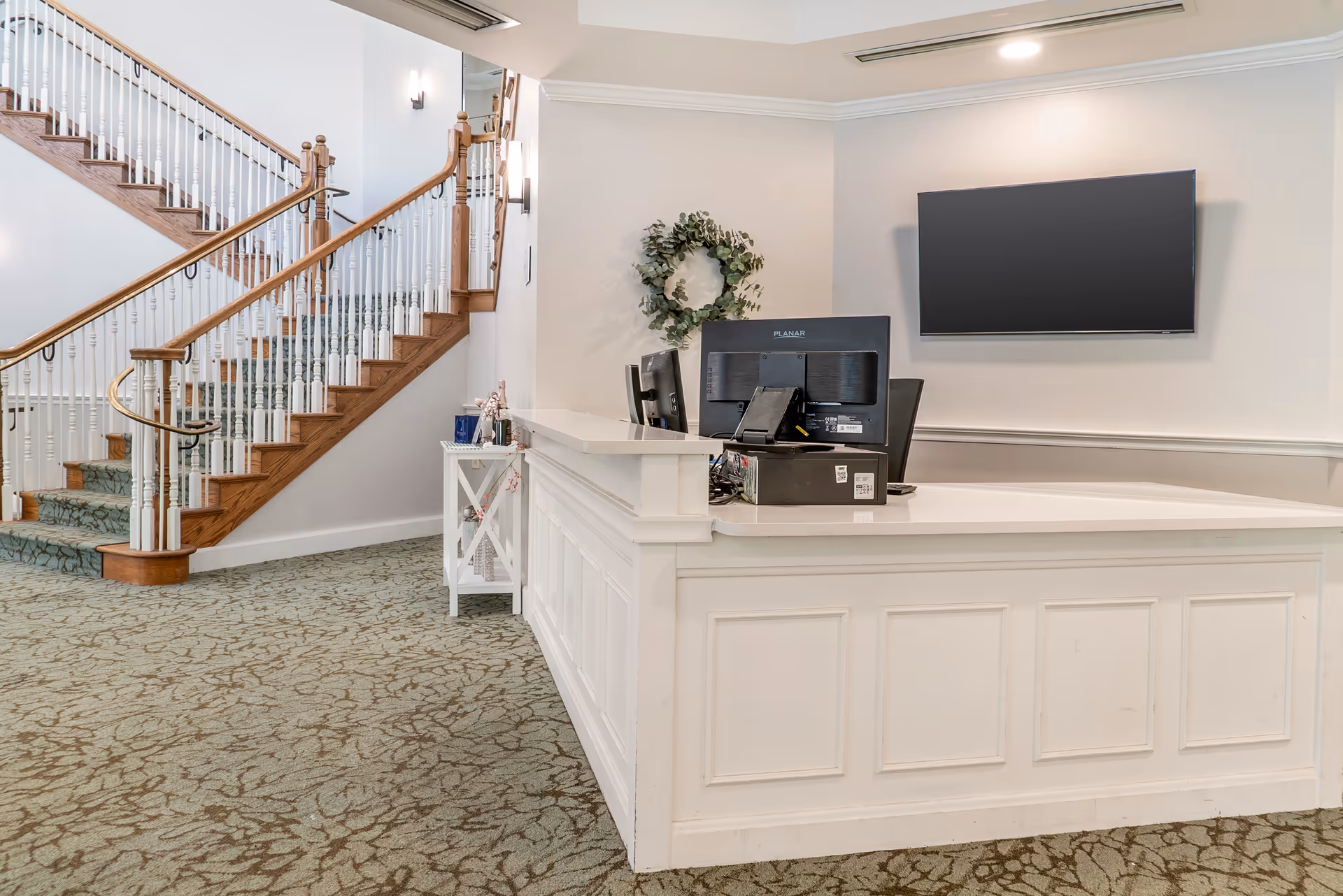Reception area with a white front desk, two computer monitors, a wall-mounted flat screen TV, a decorative wreath on the wall, and a staircase with wooden handrails and white balusters in the background.