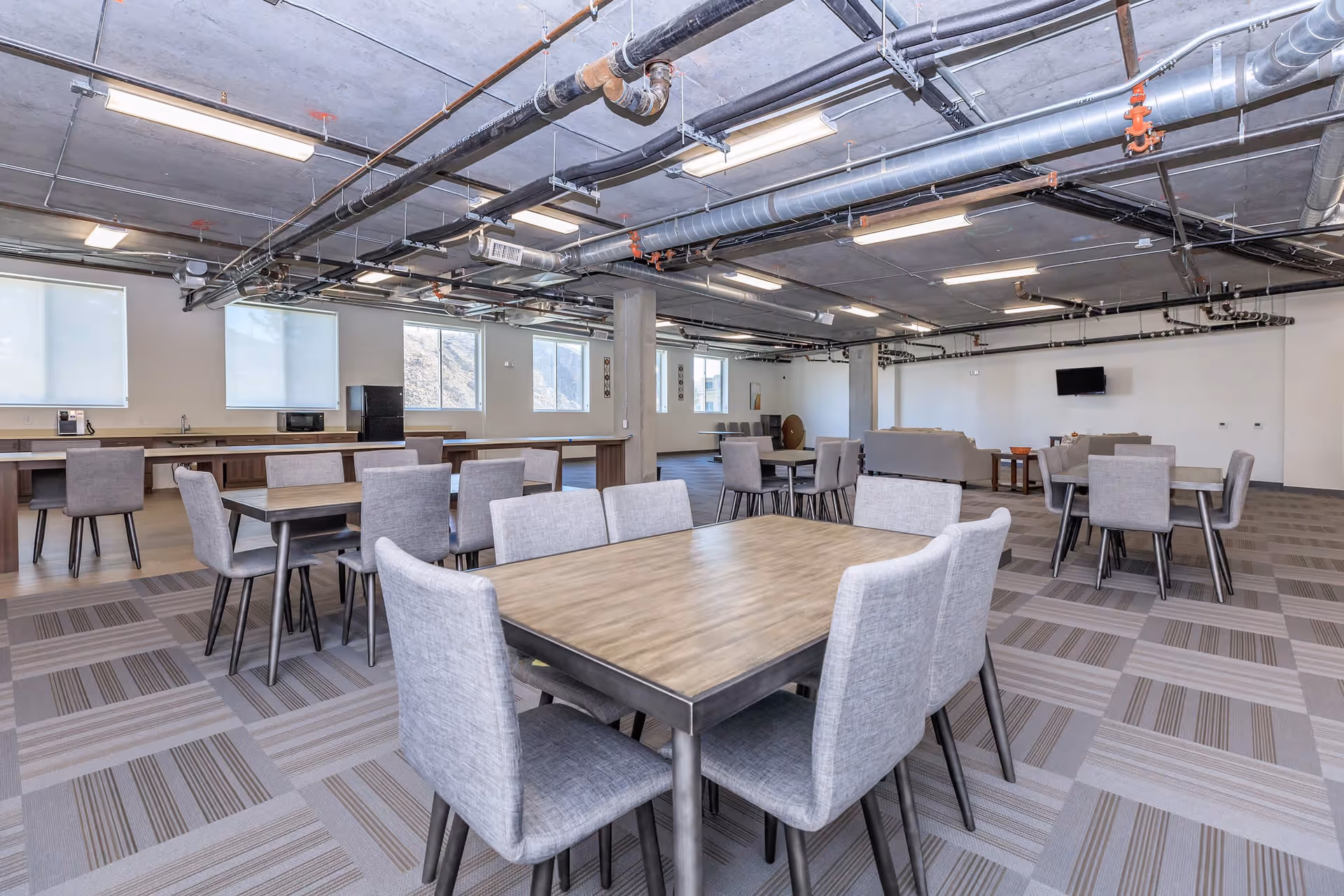 Spacious communal dining and lounge area with multiple tables and gray upholstered chairs, an exposed ceiling with visible ducts, and a kitchenette along the back wall.