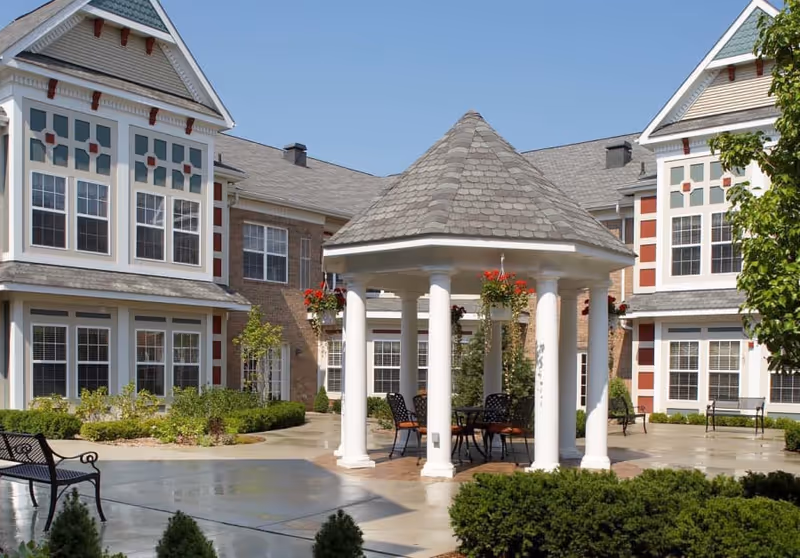 Sunny courtyard featuring a white columned gazebo with tables and chairs surrounded by landscaping and the multi-story senior living building.