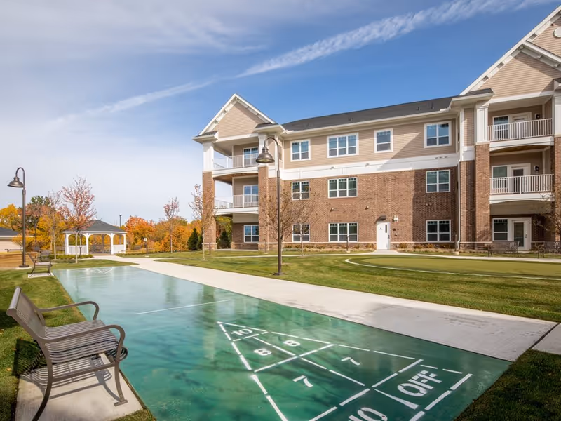 Outdoor area of a senior living facility with a shuffleboard court, benches, a gazebo, and a three-story building with balconies in the background under a partly cloudy sky.