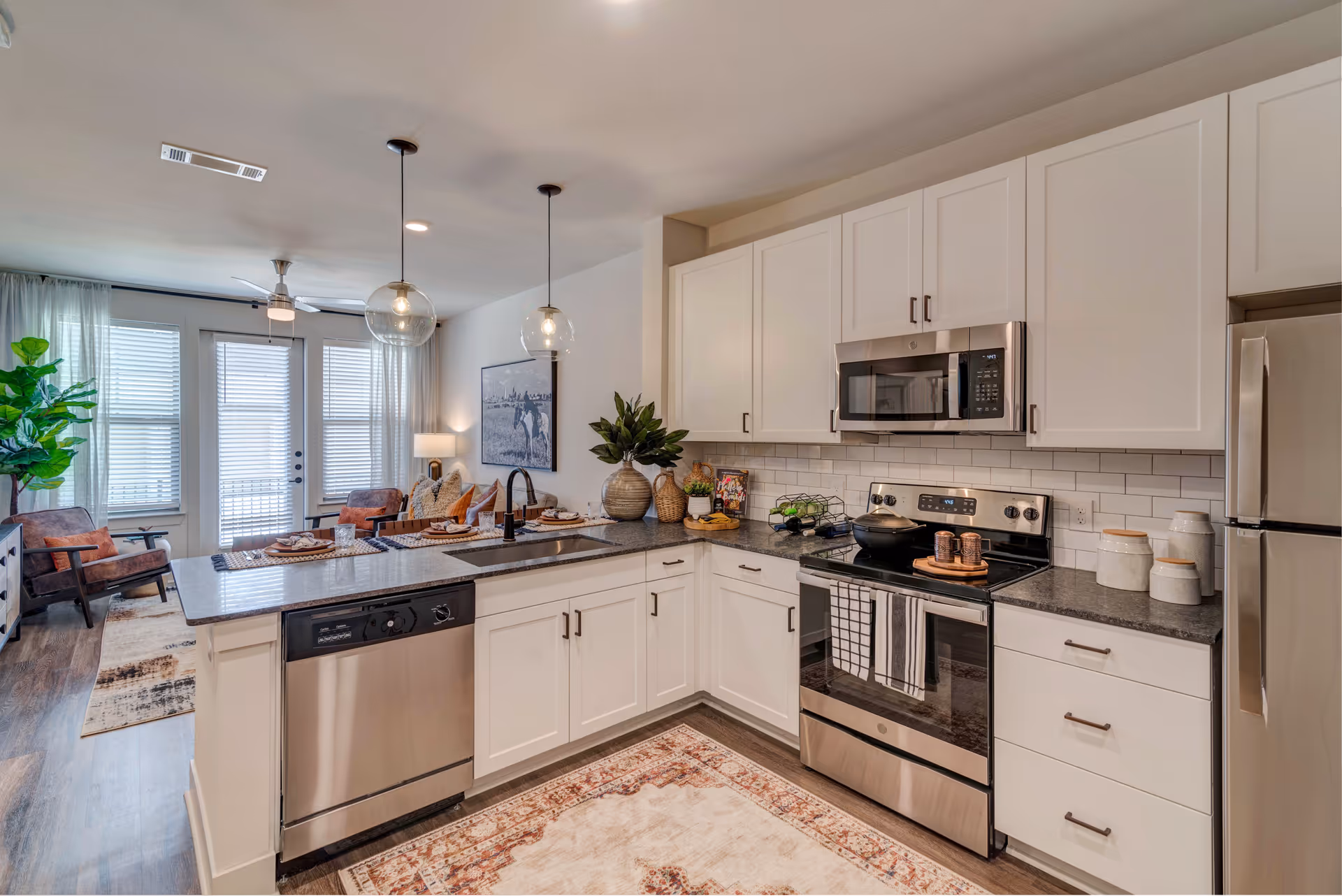 Modern kitchen with white cabinets, stainless steel appliances including a dishwasher, stove, microwave, and refrigerator. The kitchen island has a sink and two pendant lights hanging above. In the background, there is a living area with chairs, a sofa, a large plant, and a wall picture. Large windows with blinds and sheer curtains let in natural light.