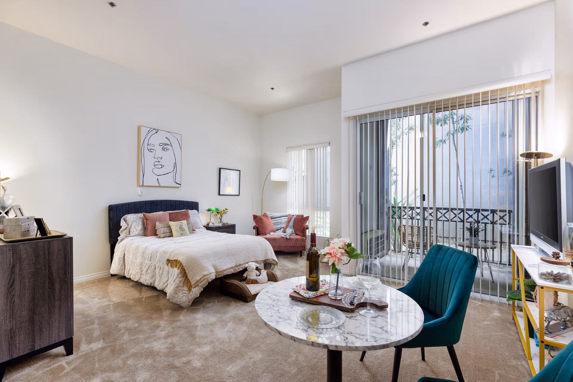 A bright and cozy bedroom with a bed featuring a dark upholstered headboard, white bedding, and several pillows. There is a small round marble table with a bottle of wine, glasses, and flowers in the foreground. A teal chair is positioned next to the table. The room has beige carpet, a dresser on the left, a TV on a yellow stand on the right, and a sliding glass door with vertical blinds leading to a small outdoor patio. The walls are white with minimalistic artwork hanging above the bed.
