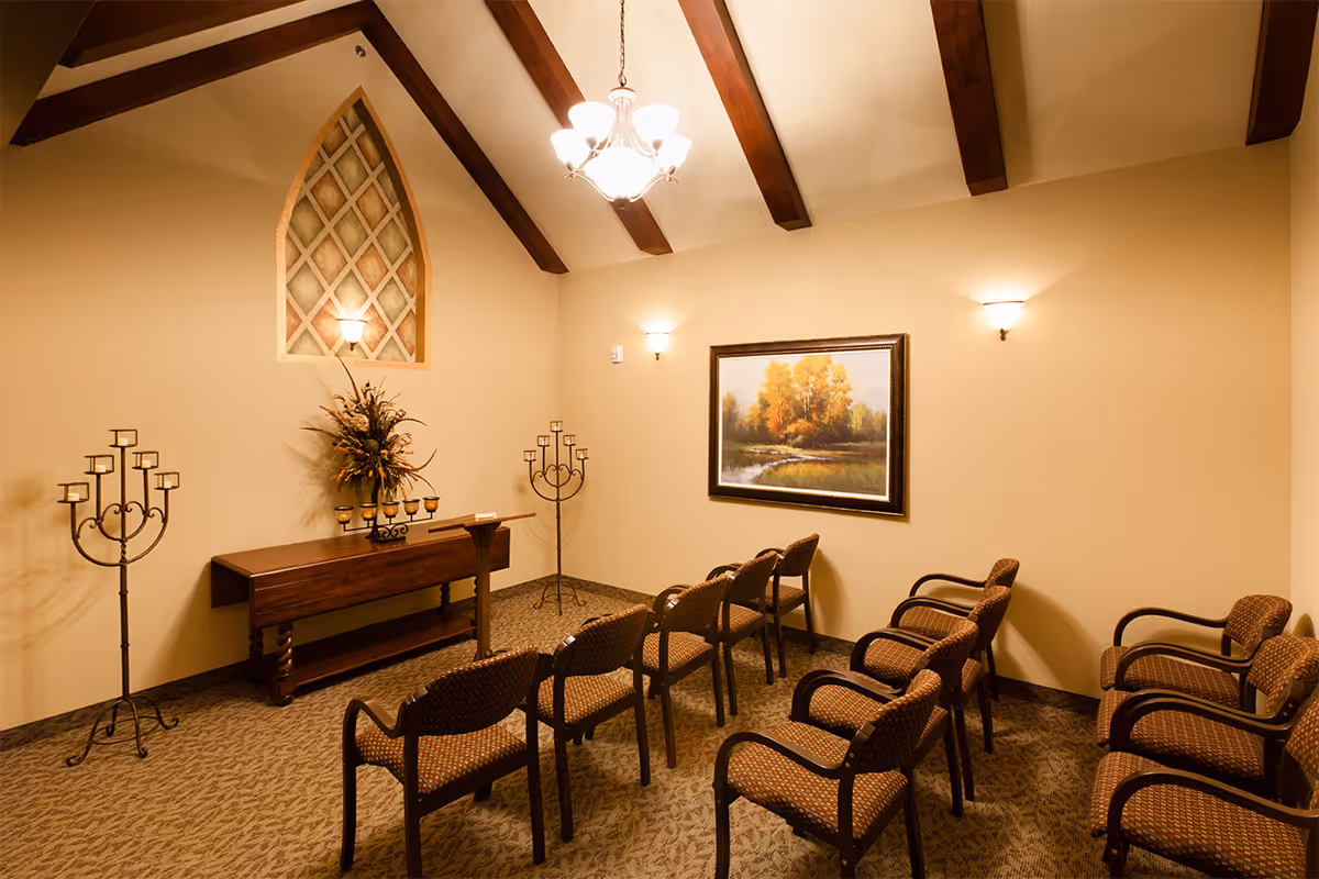Small chapel-like room with rows of padded chairs facing a table, decorative candle holders, a floral arrangement, and a framed landscape under exposed wooden ceiling beams.