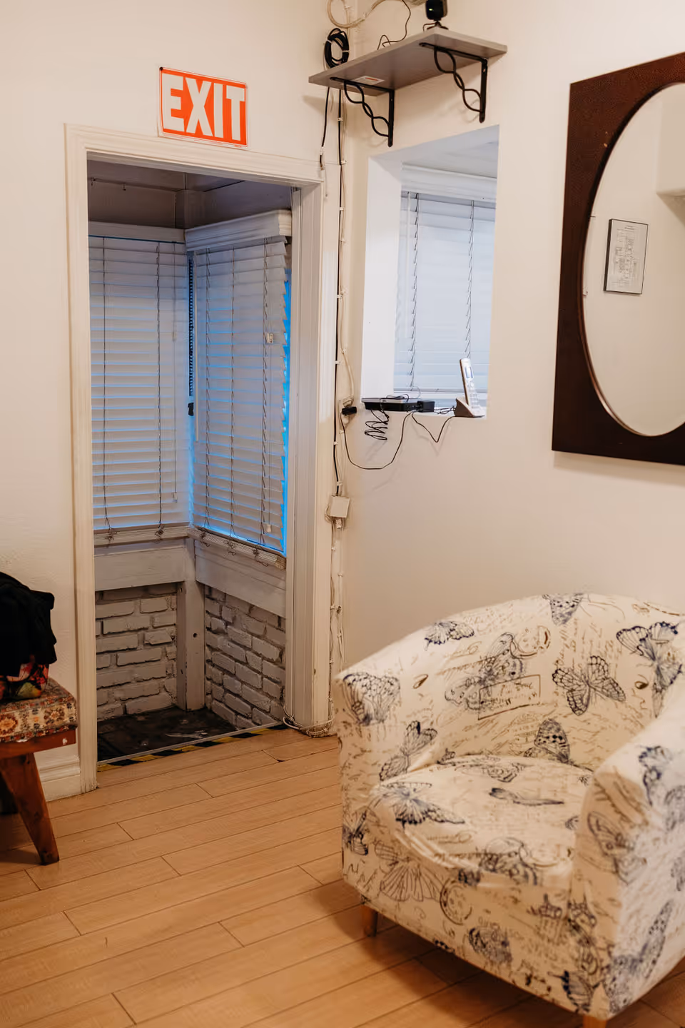 Interior room with light wooden flooring, a white armchair with butterfly and script patterns, a small wooden bench with a floral cushion, a window with closed blinds, a wall-mounted mirror, and an exit door with an illuminated red EXIT sign above it.