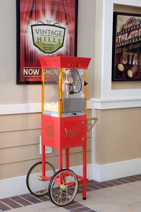 A vintage-style red popcorn machine on wheels placed indoors against a beige wall with white trim. Behind it, there are two framed posters on the wall, one advertising Vintage Hills of Indianola retirement community and the other with a theatre theme.