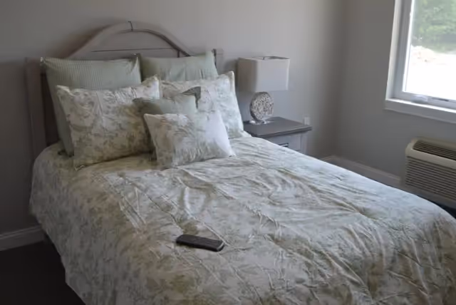 A small bedroom with a neatly made bed covered in floral bedding, a nightstand with a lamp, a window, and an air conditioning unit.