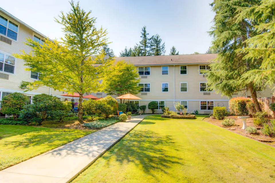 A sunny outdoor courtyard area at Bridgeport Place Assisted Living featuring a well-maintained lawn, a concrete walkway, several trees and bushes, and a beige multi-story building in the background. There are orange umbrellas providing shade over seating areas.