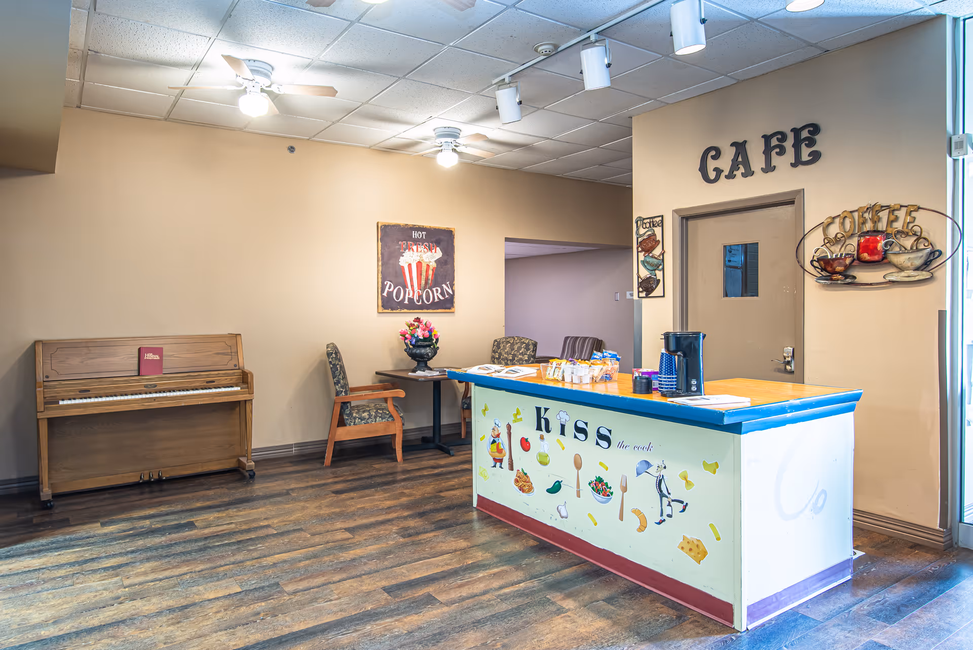 Interior cafe area with a colorful service counter, a 'CAFE' sign on the wall, a piano and seating.