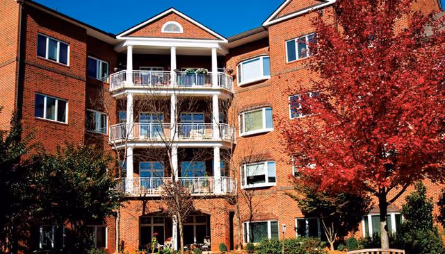 Exterior view of a multi-story red brick building with white balconies and columns, surrounded by trees including one with vibrant red autumn leaves.
