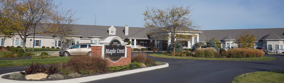 Exterior view of Maple Crest Senior Living Village showing a large single-story building with a gray roof, white walls, and multiple windows. There is a brick sign in the foreground with the facility name, surrounded by landscaped bushes and trees. A paved driveway curves around the front of the building under a clear blue sky.